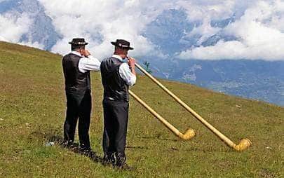 Zwei Männer in traditioneller Kleidung spielen lange Alphörner auf einer Bergwiese, im Artikel B&W Diamond Freitag.